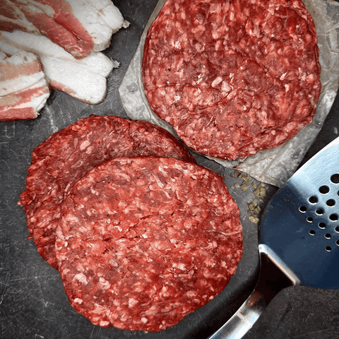 Raw hamburger and bacon patties on a pan with bacon strips and a spatula. REP Provisions